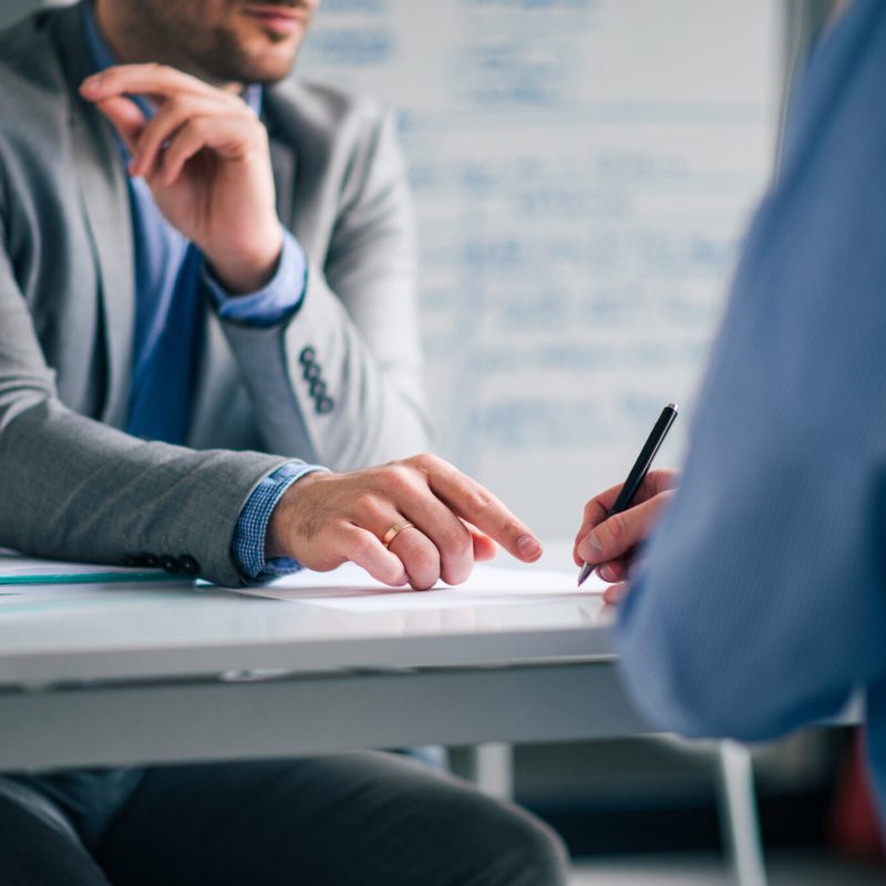 Low angle image of two men in formal wear sitting at the desk and and signing agreement.